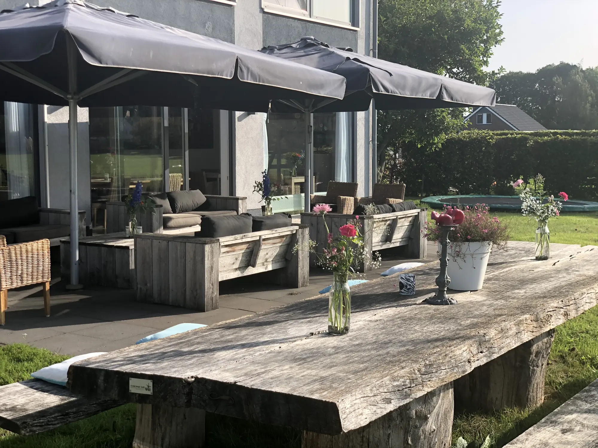 Het terras: een lange verweerde houten tafel met banken, parasols open, bloemen in vazen en de glazen pui van het huis erachter.